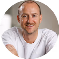 Smiling bald man with light stubble wearing a white shirt, sitting indoors with arms crossed.