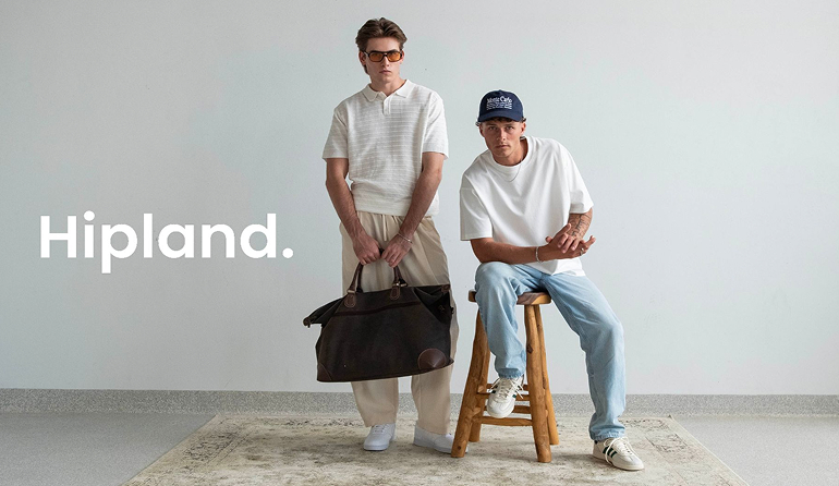 Two young men posing against a plain wall with a Hipland logo; one stands holding a large brown bag, the other sits on a wooden stool wearing a cap and casual clothes.