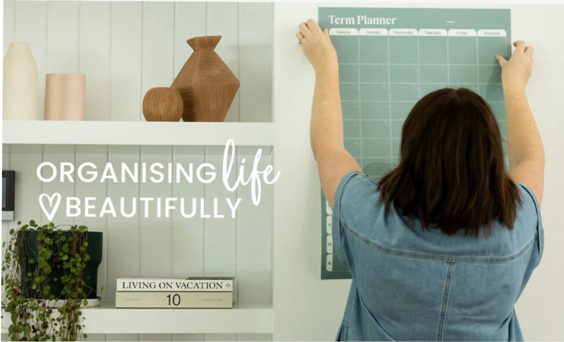 Person in denim jacket hanging a teal term planner on a white wall next to a white shelf with wooden decor, a plant, and books.