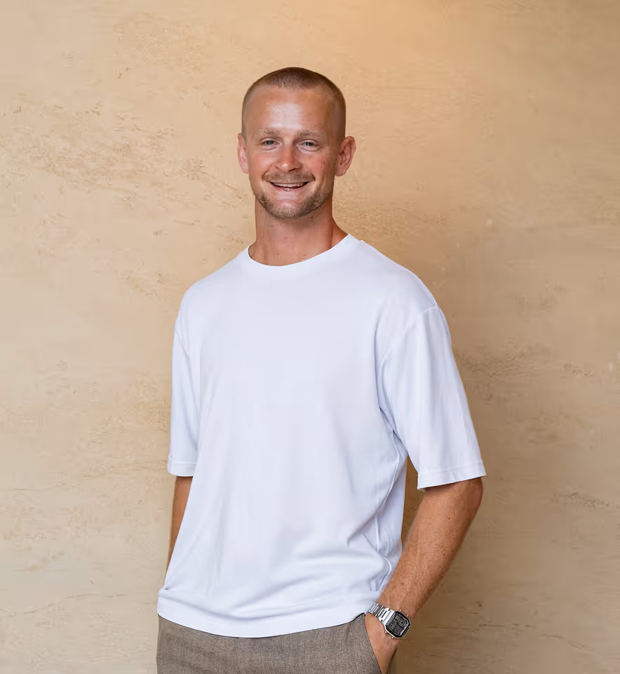 Smiling man with short hair wearing a white t-shirt and a watch, standing against a beige textured wall.