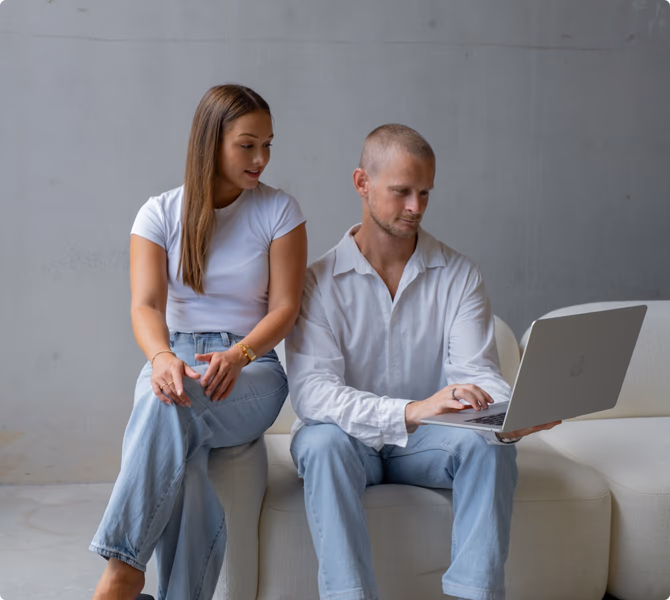 A man and woman sitting on a sofa, the man is holding and looking at a laptop while the woman looks at the laptop screen.