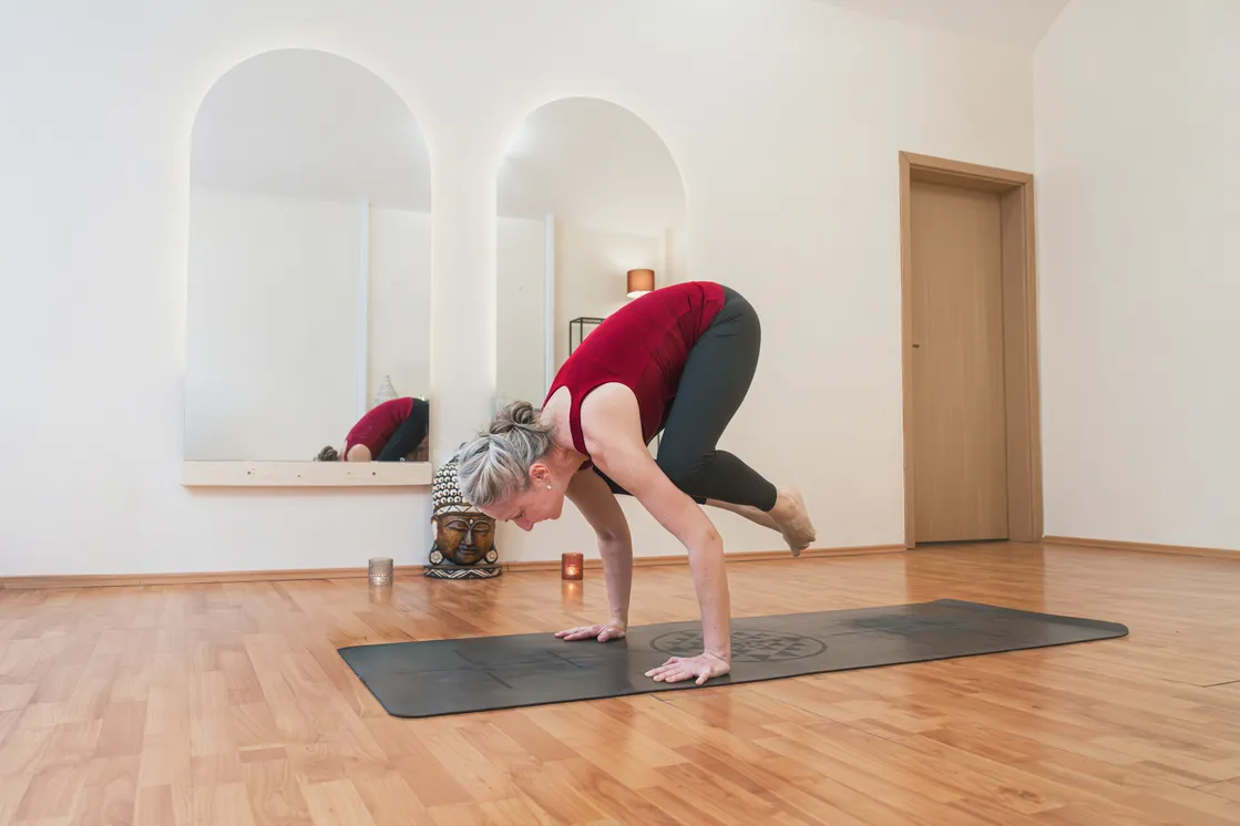 Žena v červenom tielku praktizuje jogu v pozícii žeriava na čiernej podložke v svetlej miestnosti s drevenou podlahou.