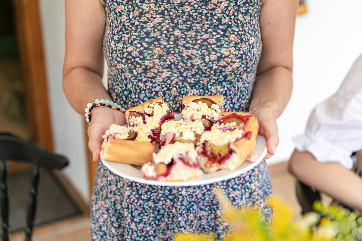 Žena v kvetinových šatách drží tanier s kúskami slivkového koláča s posýpkou.