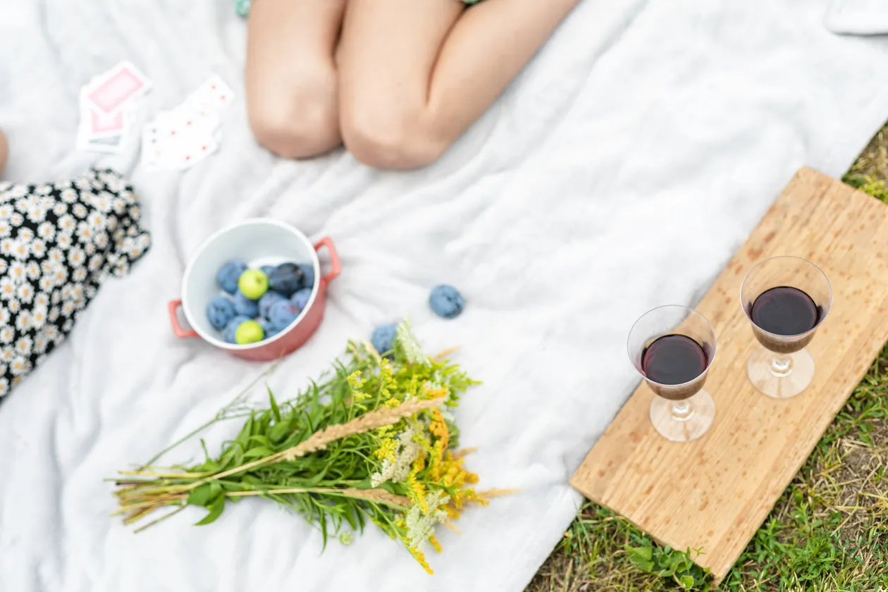 Pikniková deka s miskou sliviek, kyticou divokých kvetov a dvoma pohármi červeného vína na drevenom podnose na tráve.
