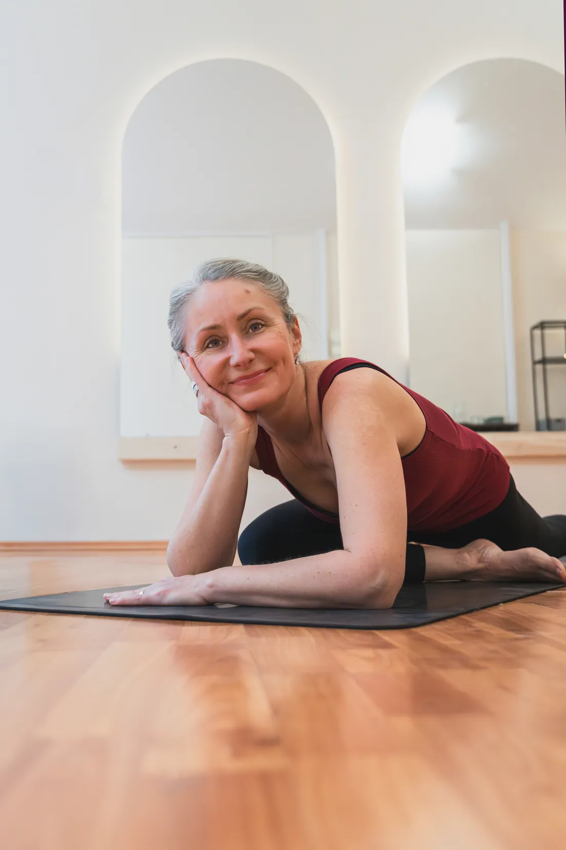 Žena cvičí jogu na čiernej podložke v svetlej miestnosti s oblúkmi na stene.