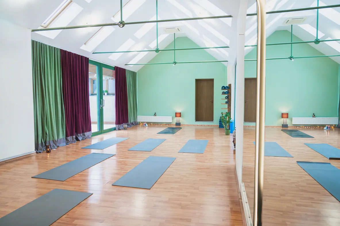 Joga štúdio so šiestimi rozloženými modrými podložkami na svetlom drevenom podlahe, veľké okná s fialovo-zelenými závesmi a zrkadlo po pravej strane.