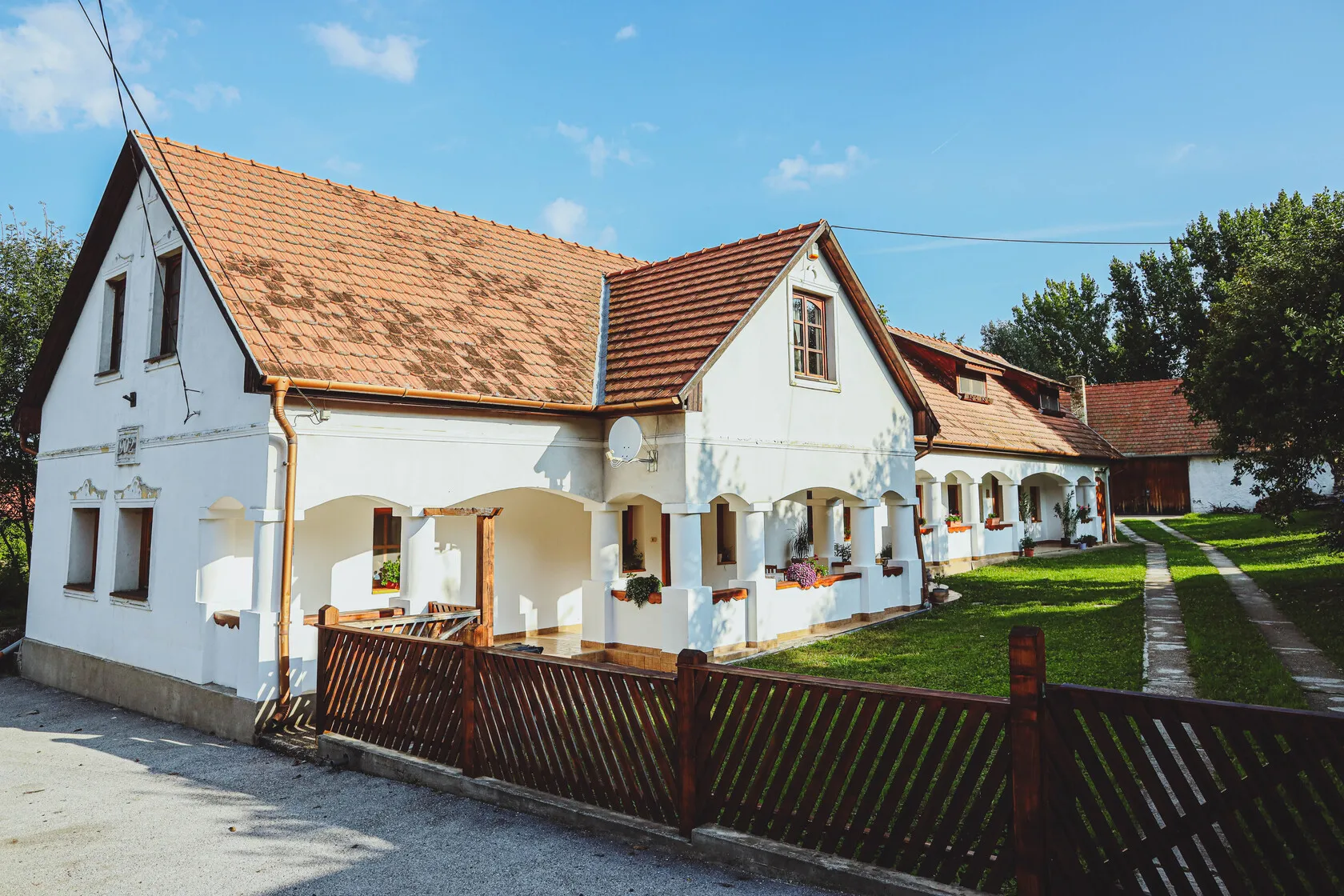 Tradičný vidiecky dom s bielymi stenami, červenou strechou a dreveným plotom pod modrou oblohou.