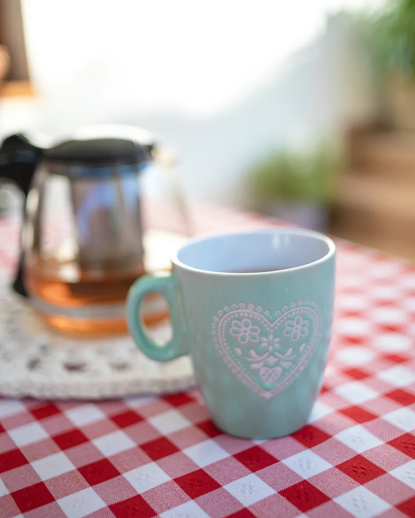 Modrý hrnček s bielym srdcom a kvetmi na červeno-bielom kockovanom obrusom s kanvicou na čaj v pozadí.
