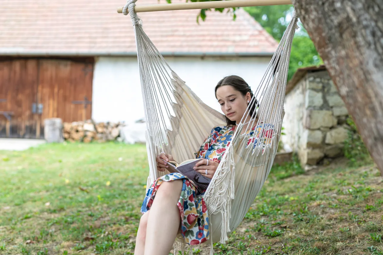 Žena čítajúca knihu sediaca v závesnom kresle na záhrade s vidieckou budovou v pozadí.