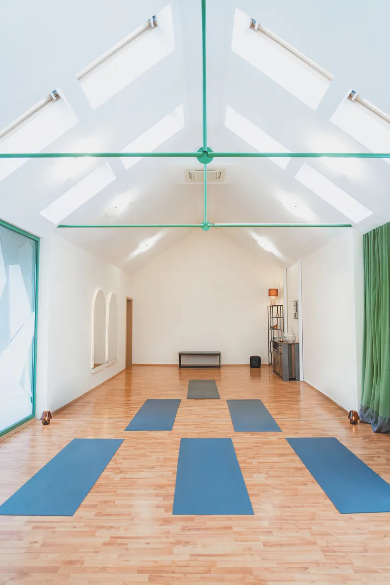 Jasná miestnosť na jogu s drevenou podlahou, modrými joga podložkami usporiadanými v radoch a zelenými nosníkmi na strope.