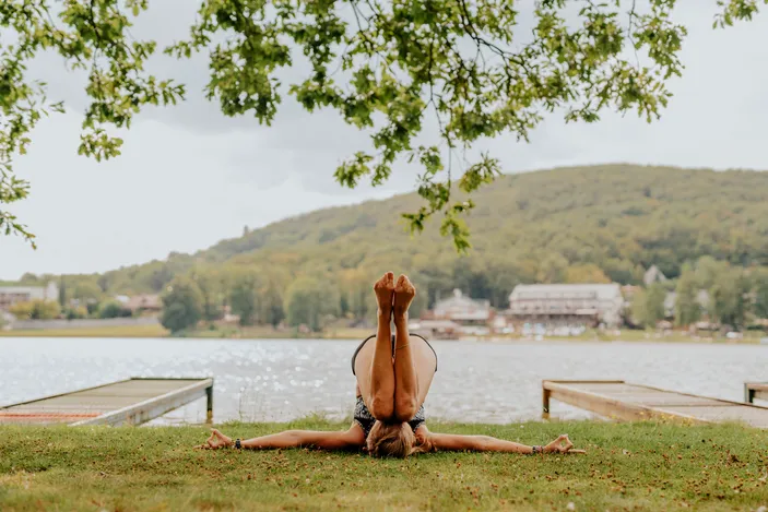 Osoba cvičiaca jógovú pozíciu na tráve pri jazere s pohorím v pozadí za slnečného dňa.