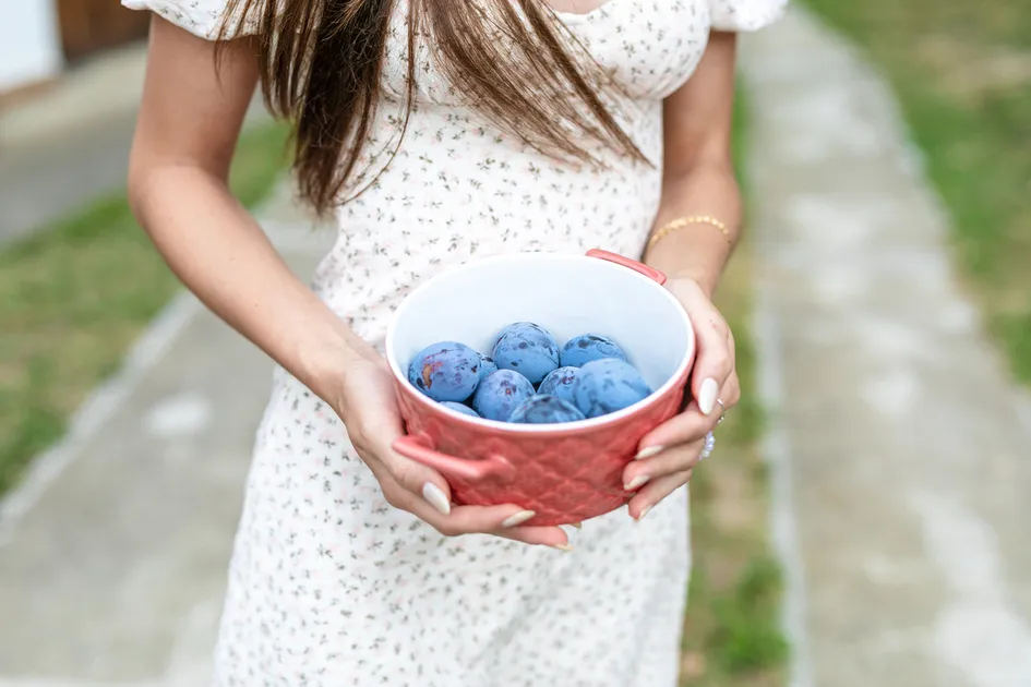 Žena v bielych šatách s kvetinovým vzorom drží červenú misku plnú modrých sliviek.