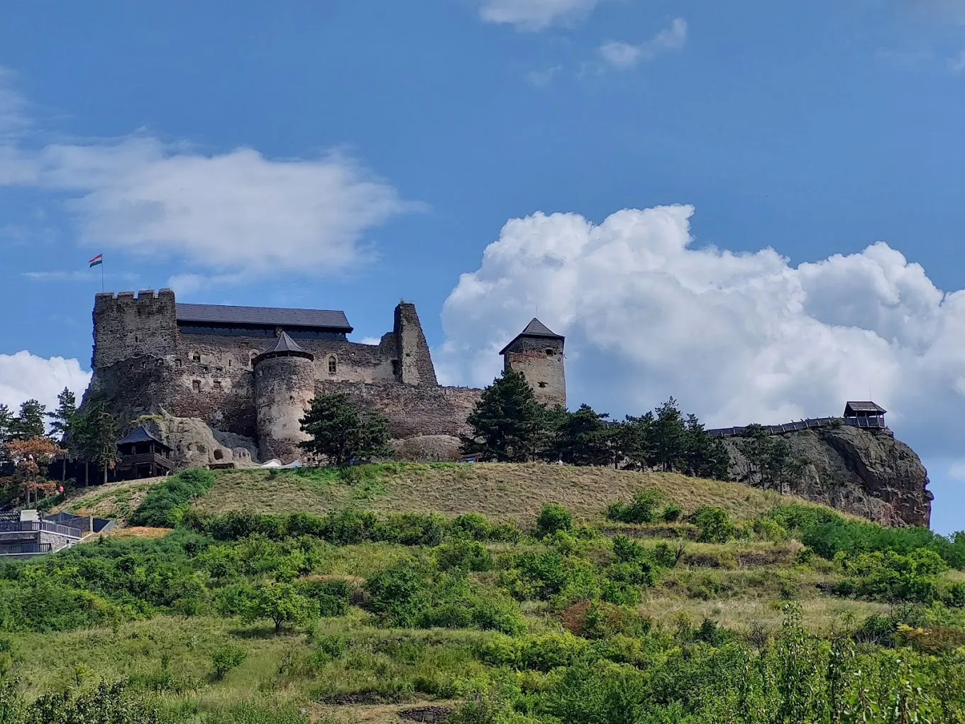 Starý kamenný hrad na kopci s malými stromami pod modrou oblohou s oblakmi.