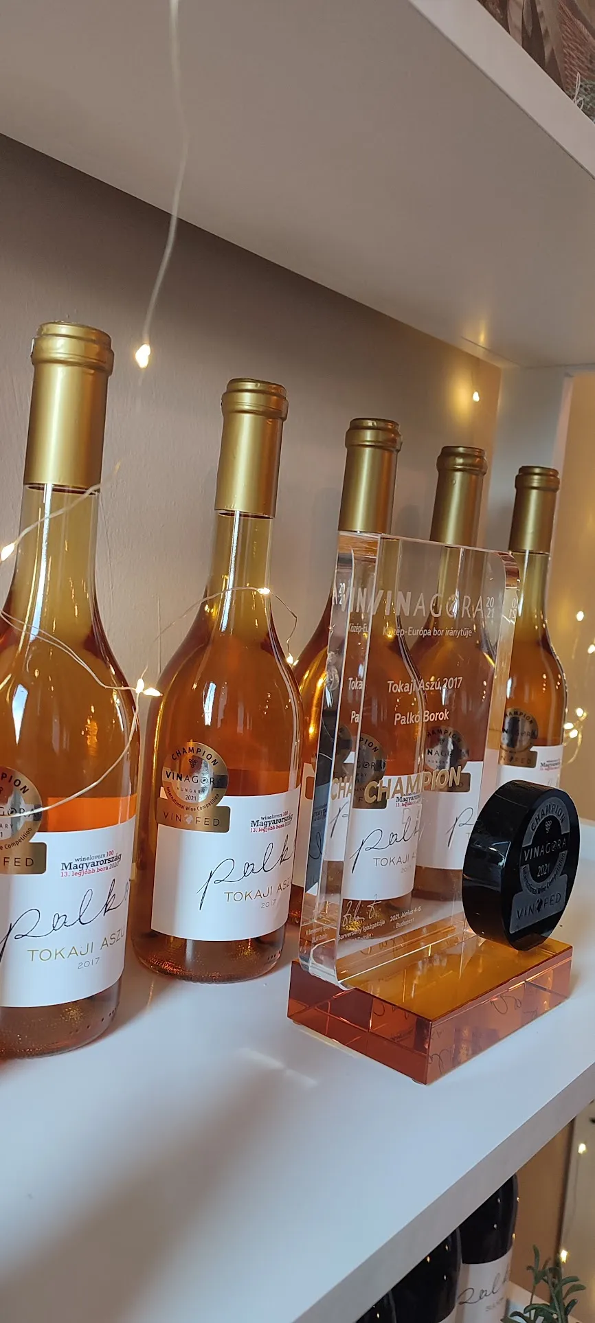 Päť fliaš vína Tokaji Aszú 2017 s medailami víťazov Vinagora a trofejami na bielom podstavci osvetlenom vianočnými svetlami.