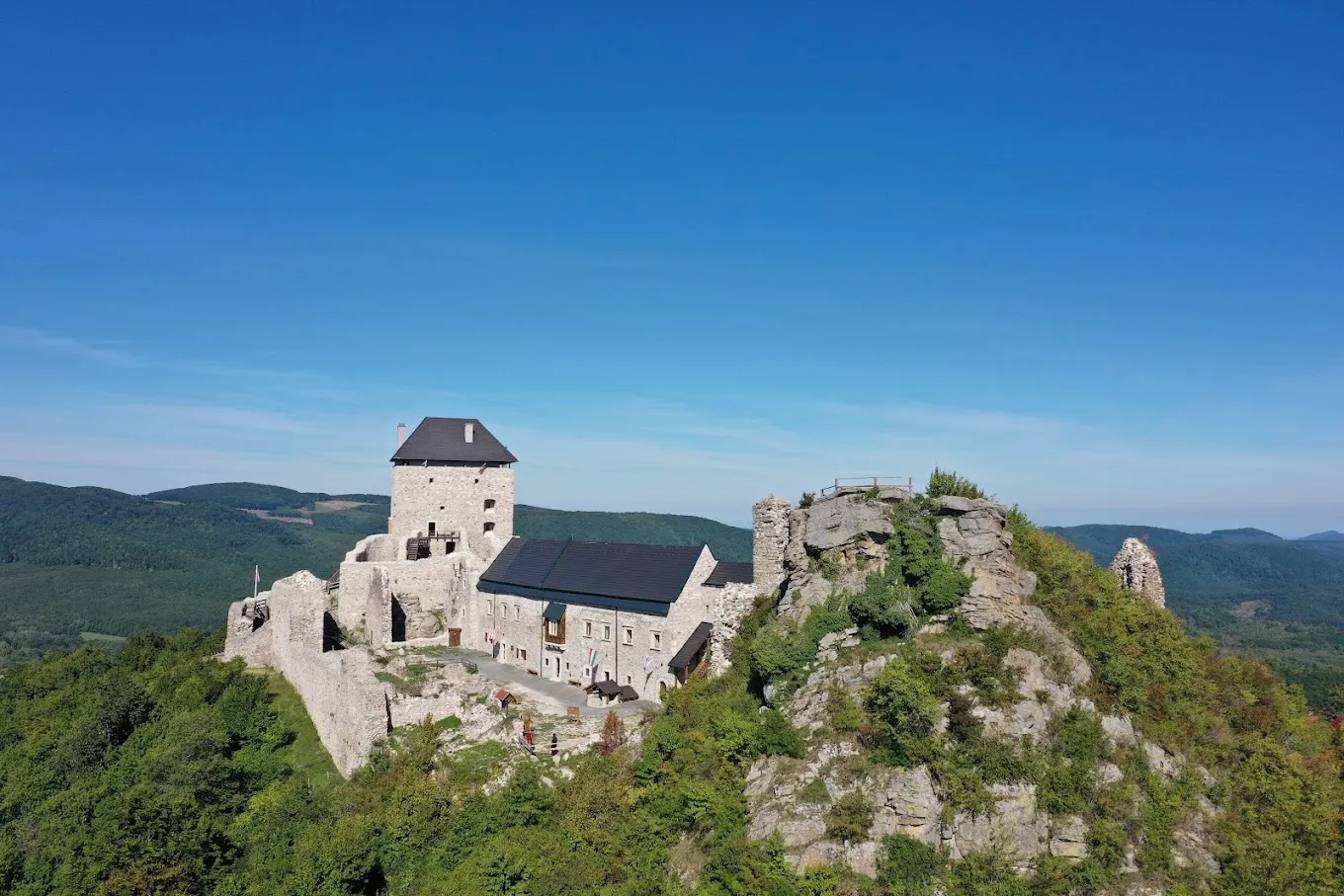 Hrad ukrytý na skalnom návrší obklopený zelenými stromami a kopcami pod jasnou modrou oblohou.