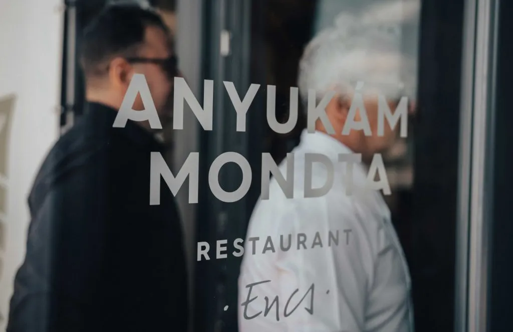 Nápis na skle reštaurácie Anyukám Mondta s rozmazanými postavami muža a kuchára v pozadí.