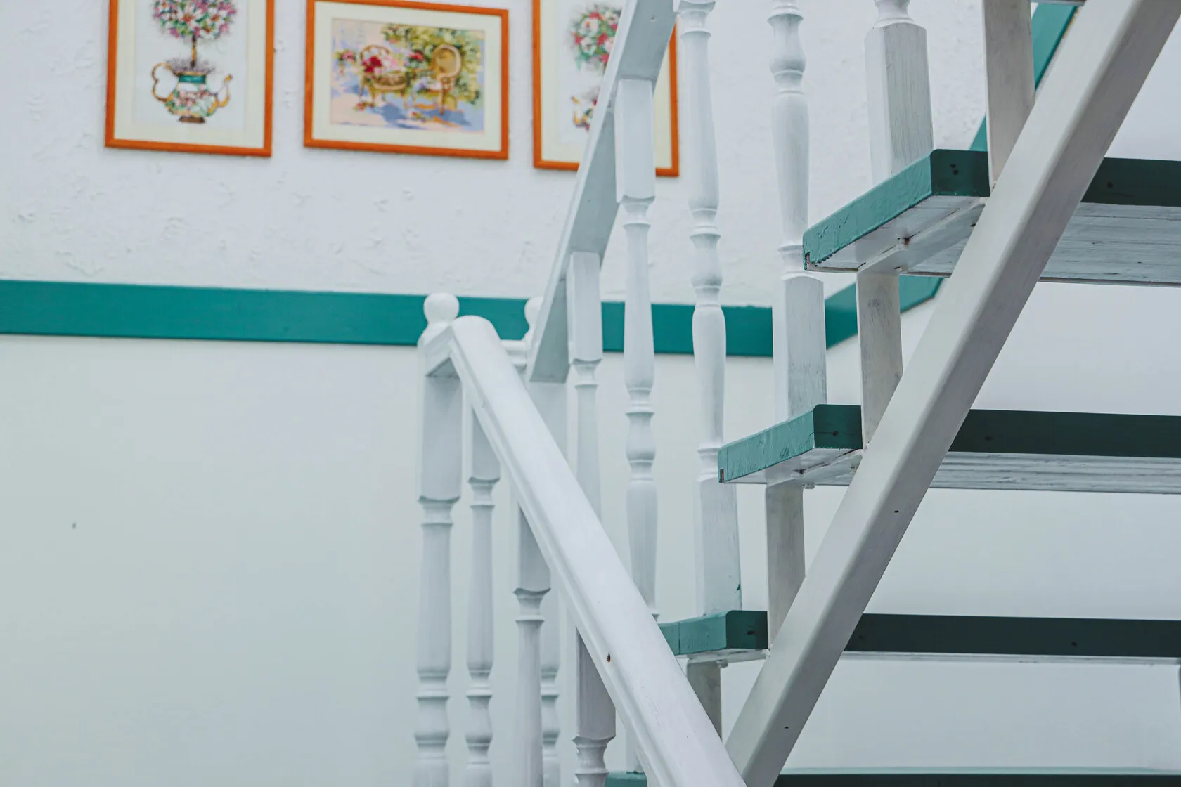 Biele drevené schodisko s mätovozelenými hranami stupňov v interiéri s bielou stenou a tromi zarámovanými maľbami nad zeleným lemom.