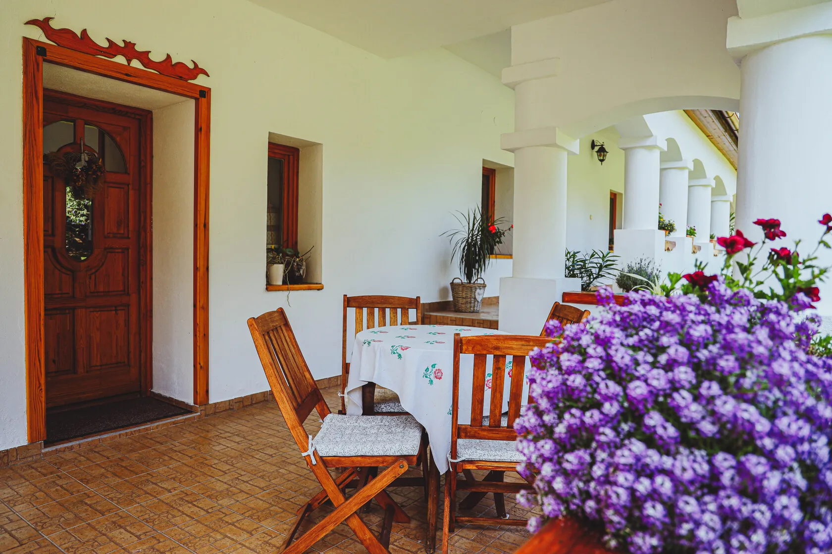 Terasa s dreveným stolom a stoličkami, okolo nich biele steny s drevenými okennými rámami a kvetináče s rastlinami.