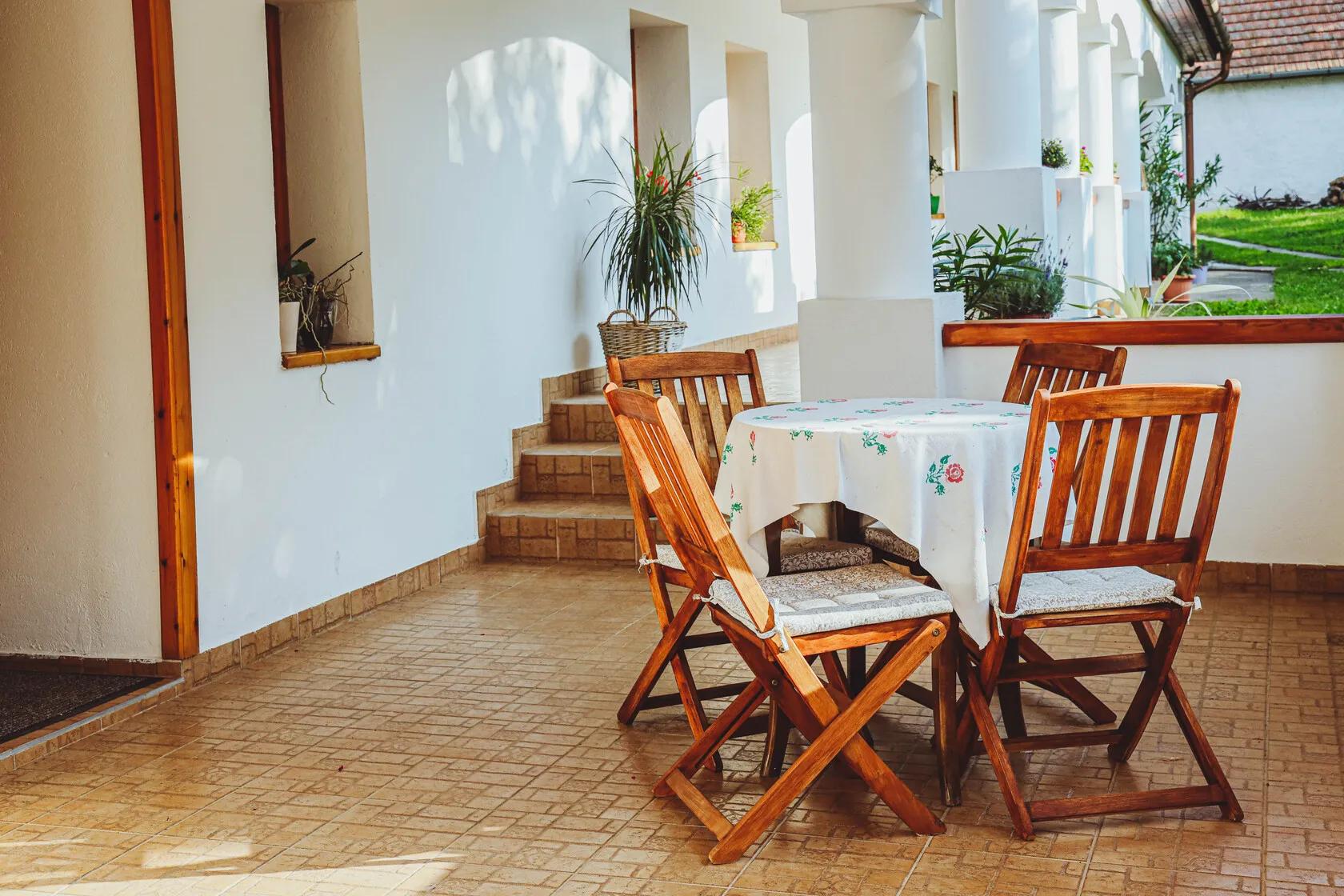 Drevený jedálenský set so štyrmi stoličkami a stolom s obrusom na presvetlenej terase s bielymi stenami a rastlinami.
