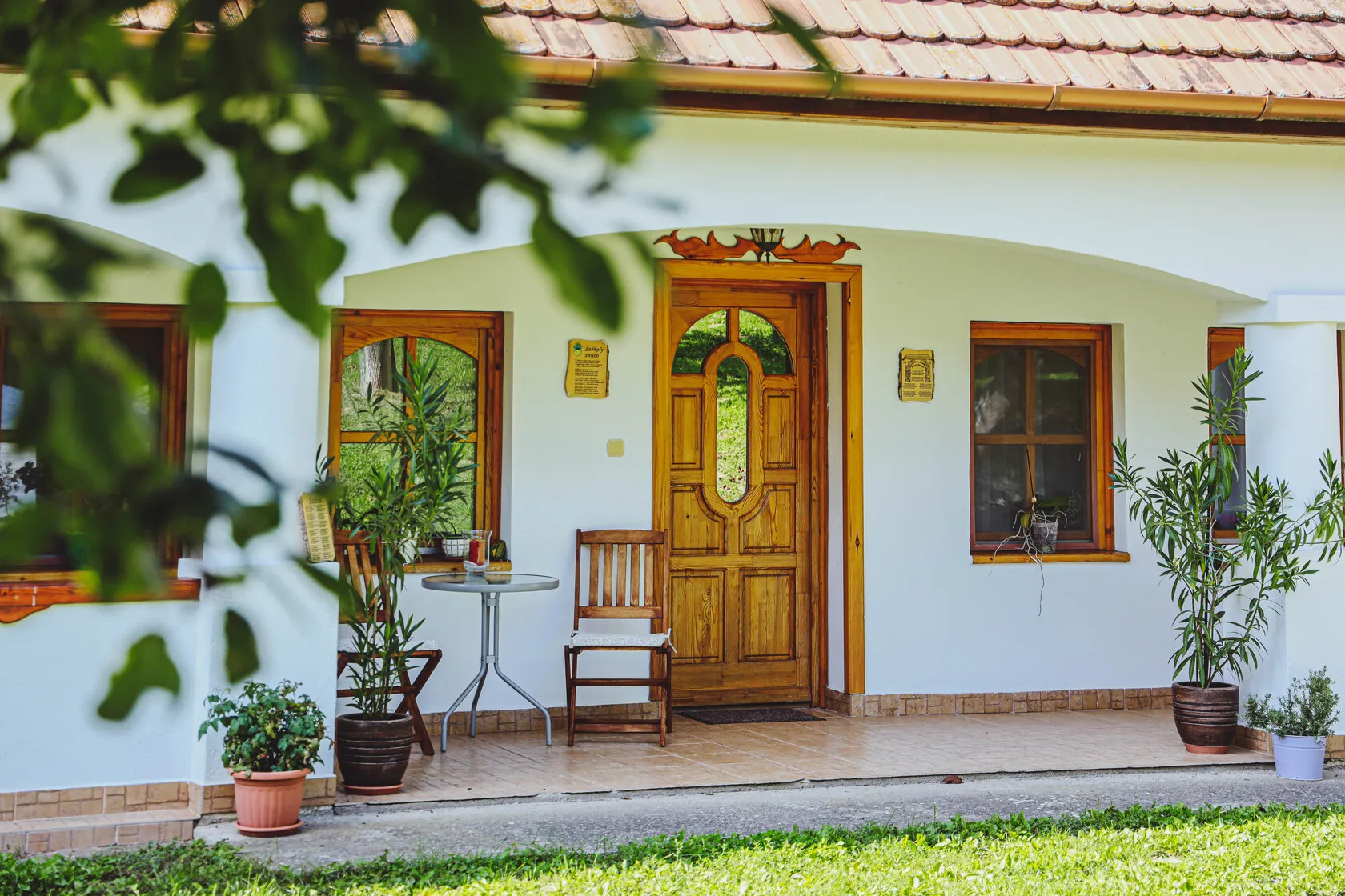 Vstup do domu s drevenými dverami a oknami, pred domom sú drevené stoličky, okrúhly stôl a kvetináče s rastlinami.