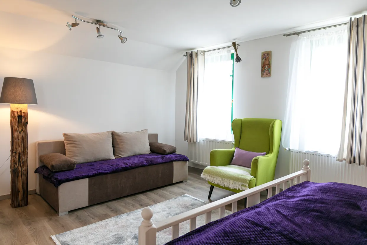 Obývacia izba s béžovou pohovkou, zeleným kreslom, lampou z dreva a dvoma oknami s bielymi závesmi.