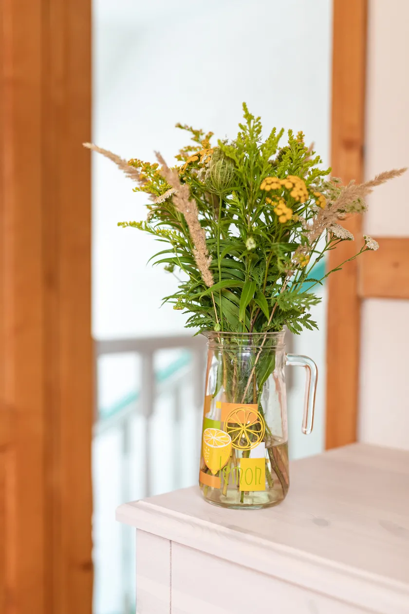 Sklenená karafa s kvetinovou dekoráciou so žltými kvetmi a zelenými listami na bielom stole.