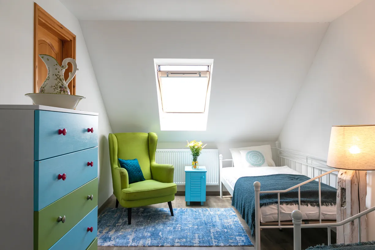 Útulná miestnosť so zeleným kreslom, modrou komodou, posteľou s modrou prikrývkou a svetlom lampou pod strešným oknom.