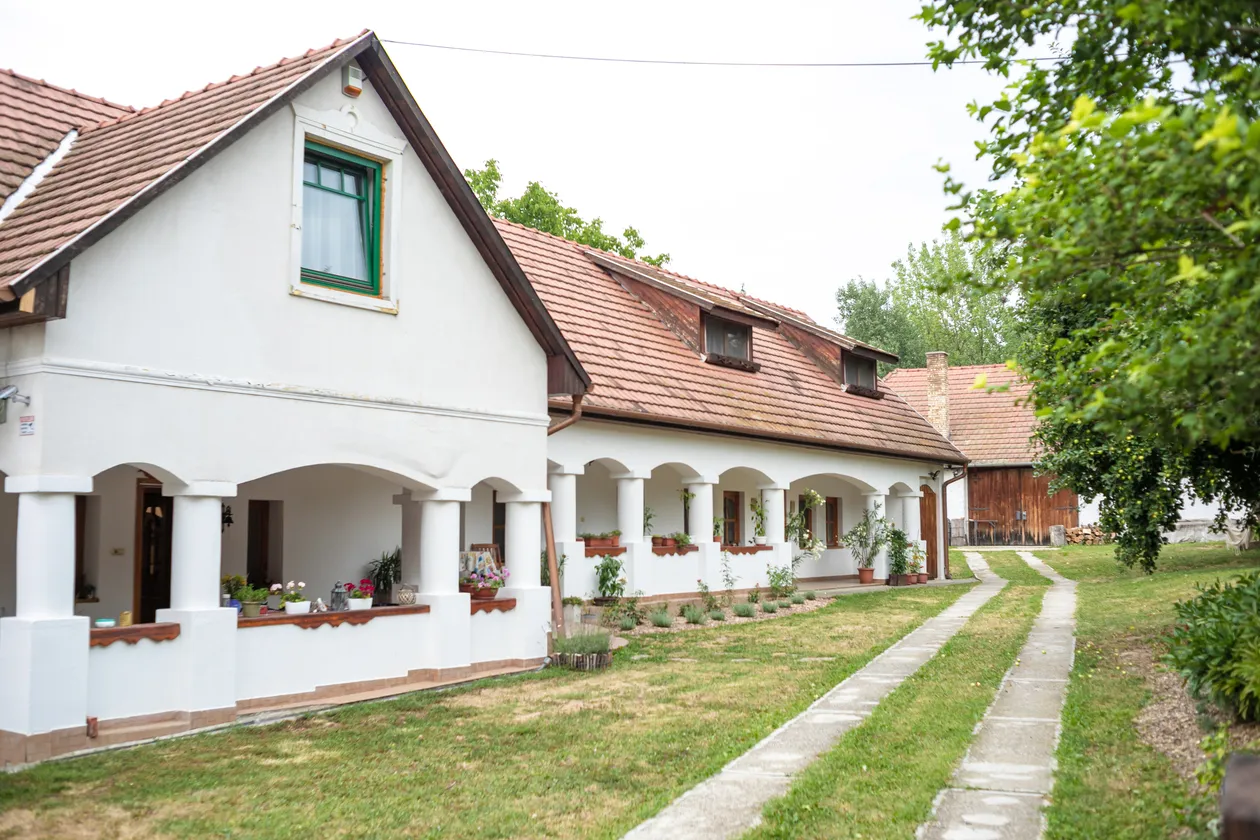 Tradičný vidiecky dom s bielymi stenami, červenou strechou a verandou lemovanou kvetmi, vedľa cesty s trávou.