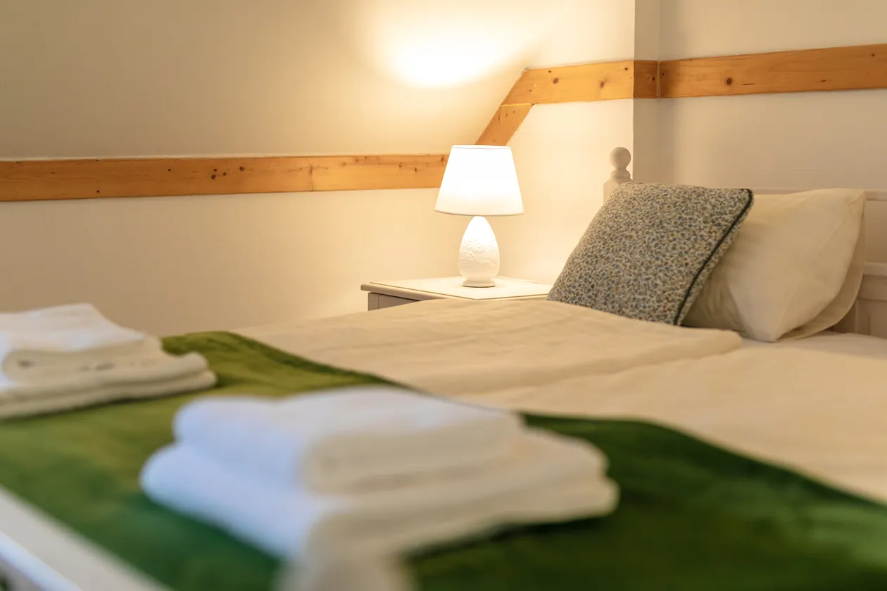 Posteľ s bielym obliečkami, dekoratívnym vankúšom, nočným stolíkom a zapnutou stolovou lampou v útulnej spálni.