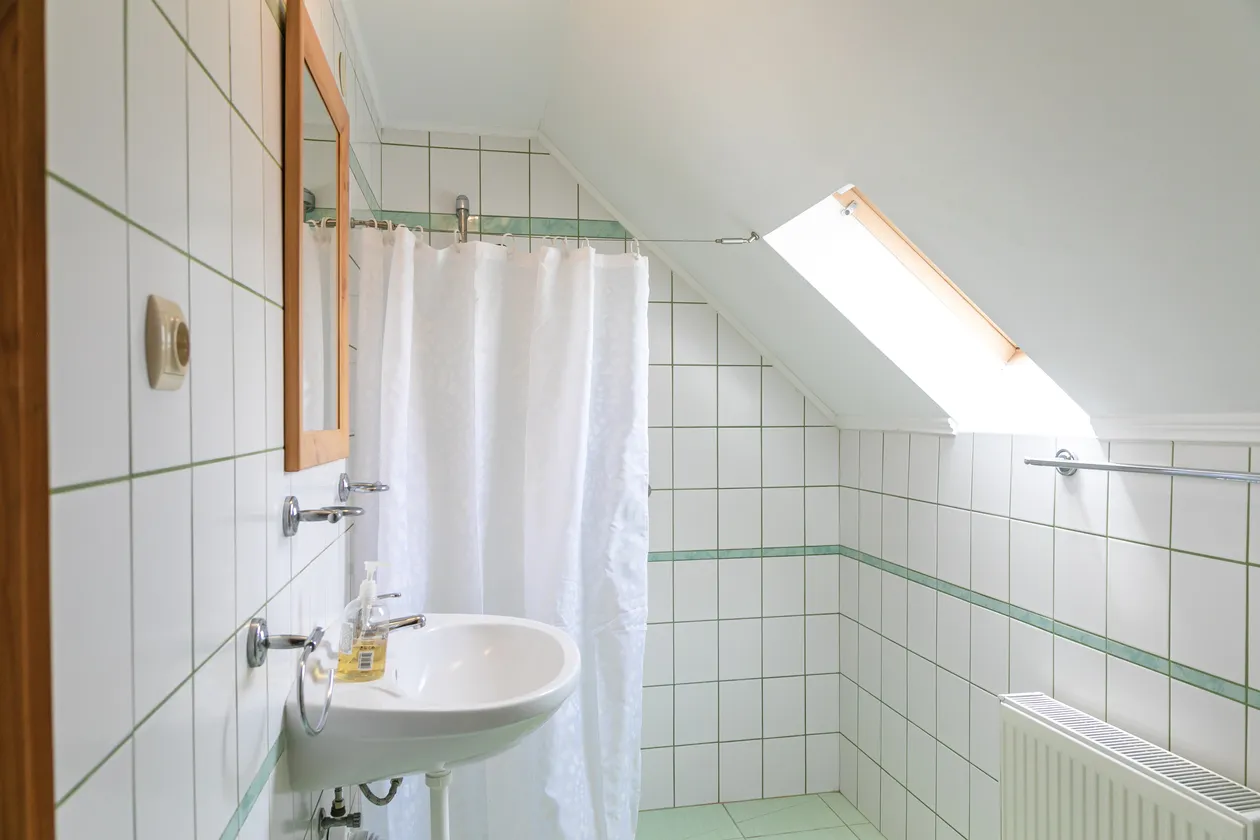 Malá biela kúpeľňa pod šikmým stropom s umývadlom, zrkadlom, sprchovacím závesom a strešným oknom.