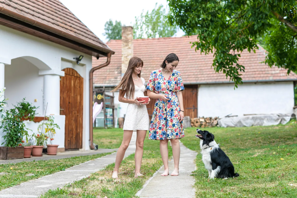 Dve mladé ženy v letných šatách stojace na chodníku pred domom, ktoré sa pozerajú na sediaceho čierno-bieleho psa na tráve.