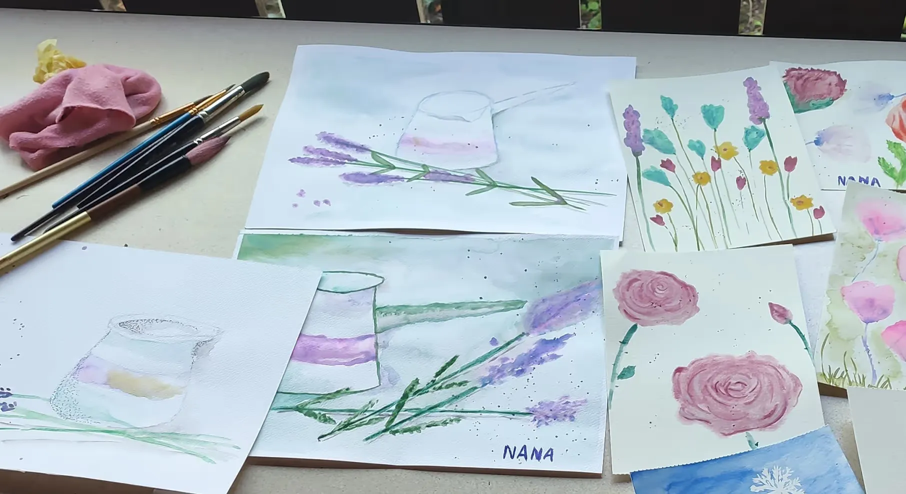 Rôzne akvarelové maľby kvetov a konvičiek s niekoľkými maliarskymi štetcami a handričkou na stole.