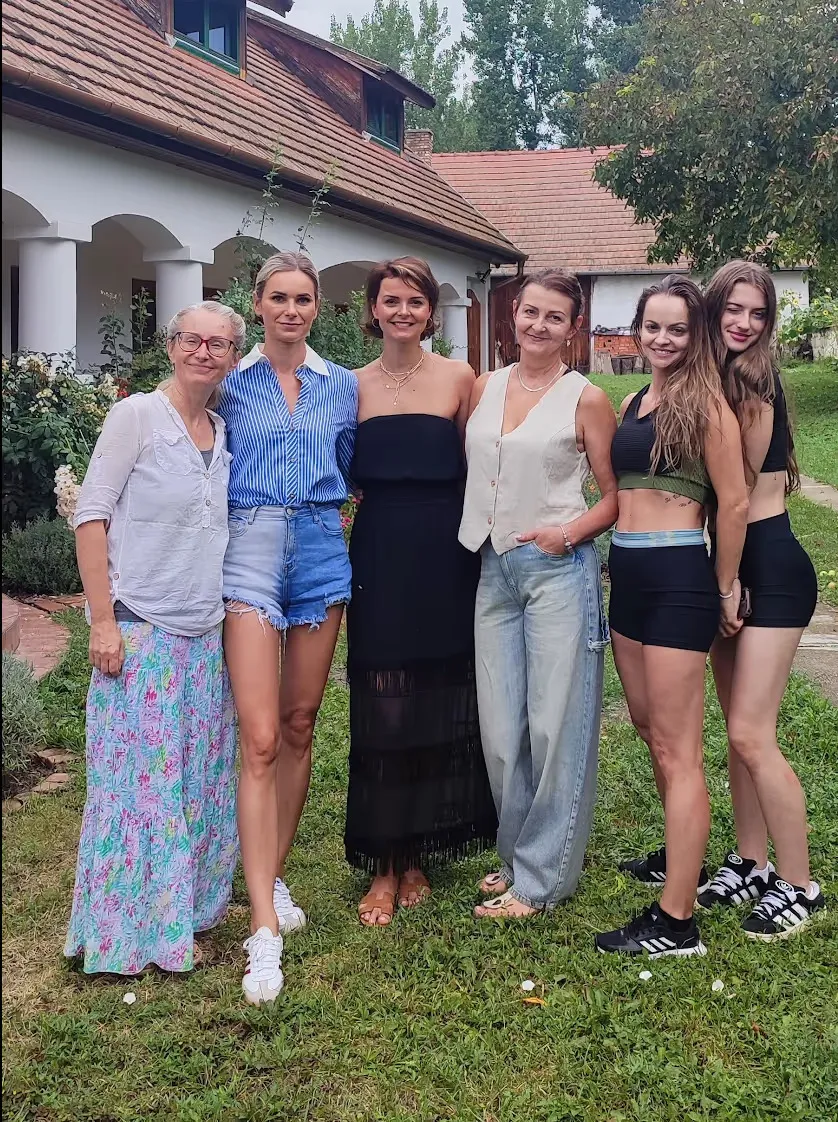 Šesť žien rôzneho veku stojí spolu vonku na trávniku pred domom s červenou strechou, niektoré sa usmievajú.