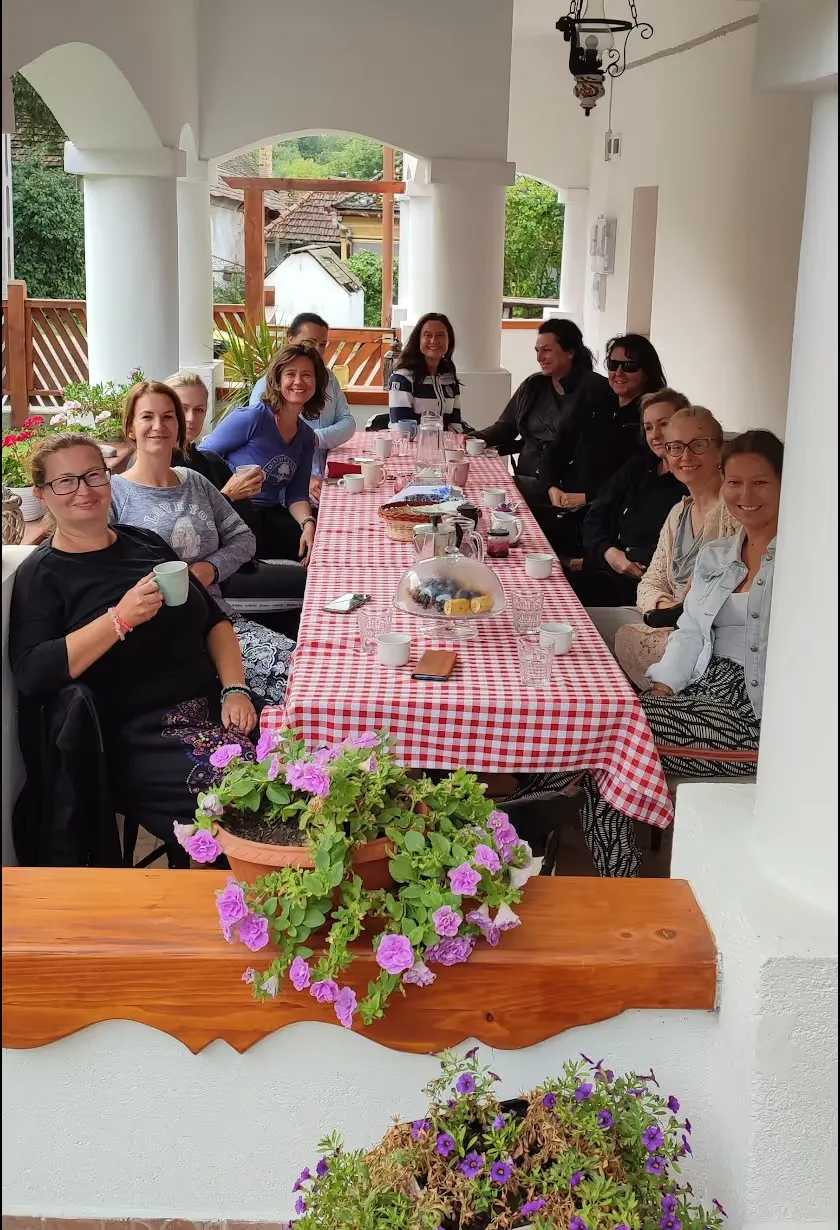 Skupina žien sedí okolo stola s červeno-bielym károvaným obrusom na krytej terase s kvetinovými nádobami v popredí.