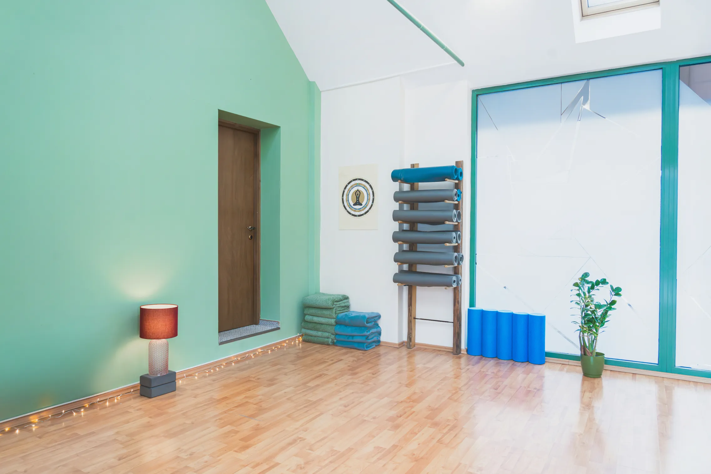 Jasná miestnosť na cvičenie s drevenou podlahou, zelenou stenou, lampou, uterákmi, penovými valcami a izbovou rastlinou pri veľkom okne.