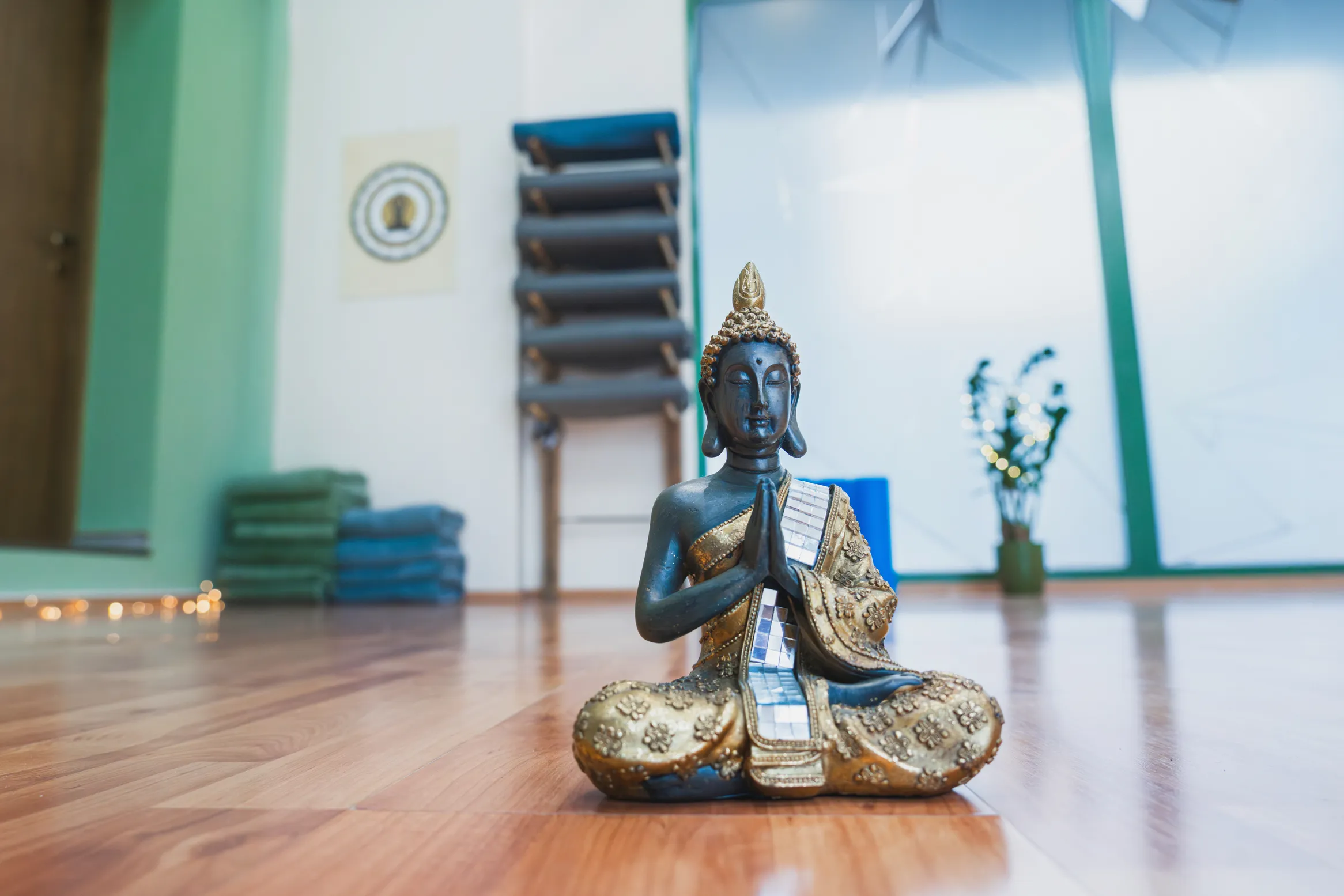 Socha sediaceho Budhu v meditačnej miestnosti s drevenou podlahou, v pozadí skladané podložky a rastlina s rozmazaným svetlom.