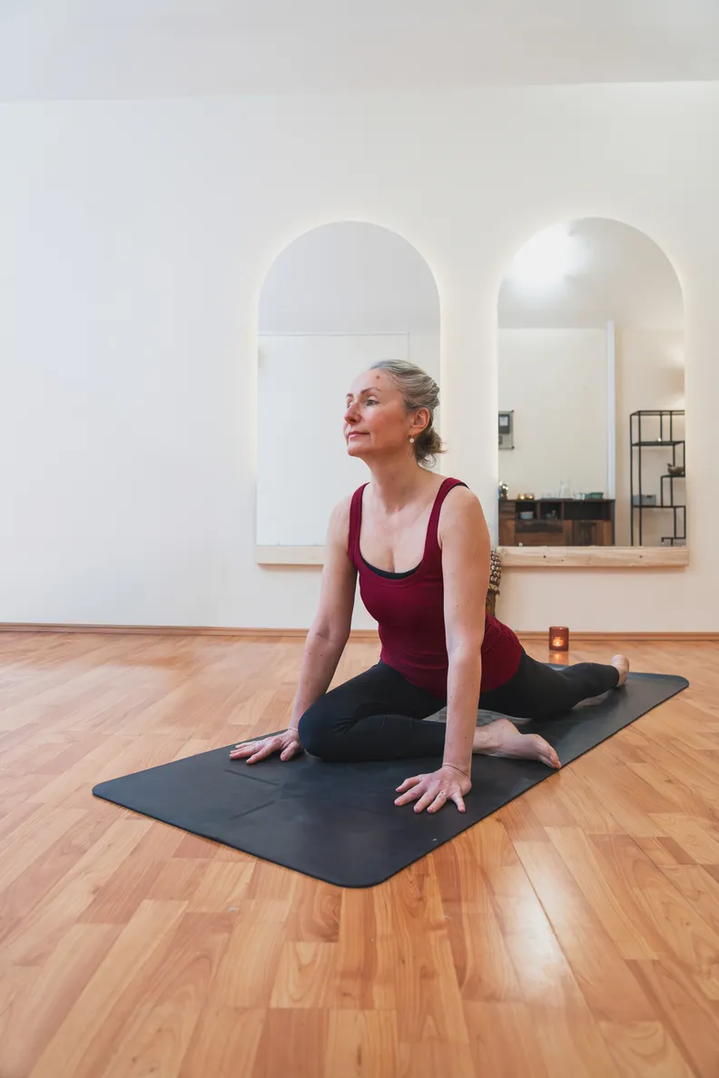 Žena cvičiaca jógu v miestnosti s umelým svetlom, sedí v póze holuba na čiernej podložke na drevenej podlahe.