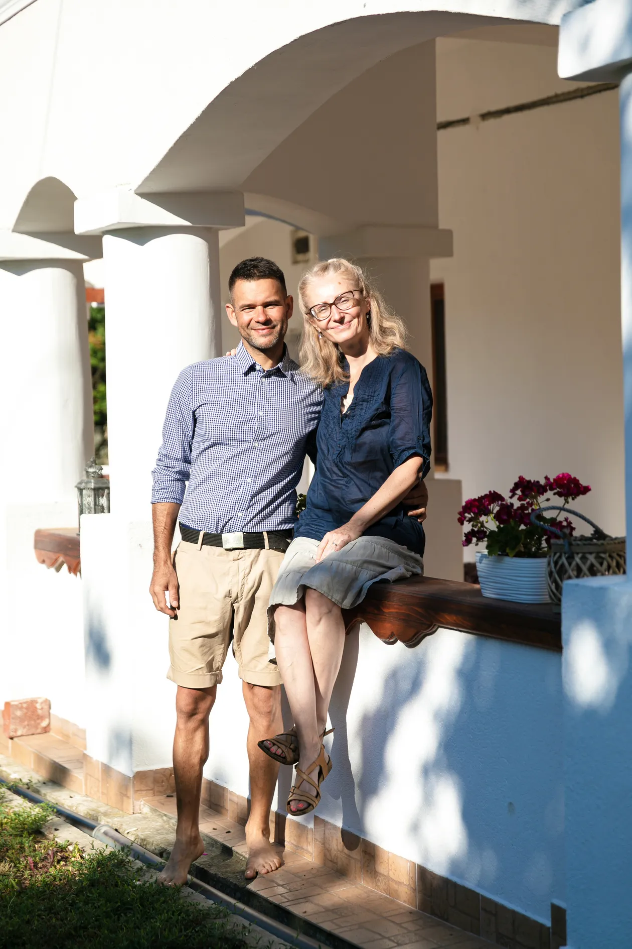 Muž v béžových šortkách a modrej košeli s károvaným vzorom stojí na boso vedľa ženy sediacej na drevenom parapete pod bielou klenbou.