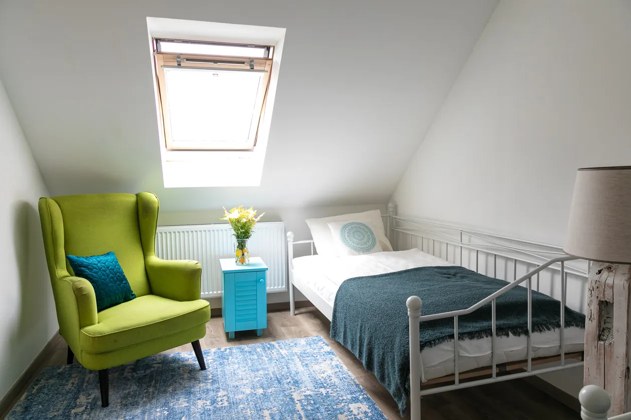 Komfortná izba so zeleným kreslom, modrým nočným stolíkom, bielou posteľou a oknom na strope.