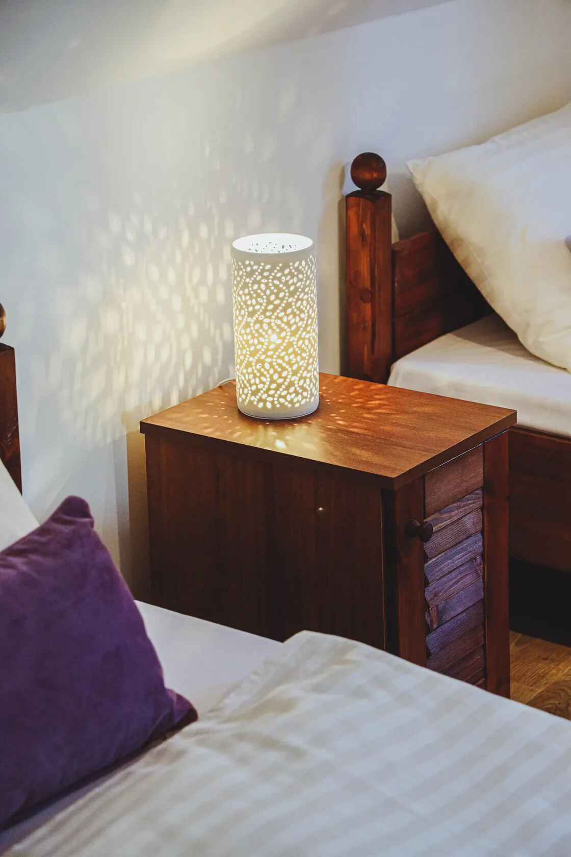 Stolná lampa s perforovaným vzorom vrhá svetelné efekty na drevený nočný stolík medzi dvoma posteľami.