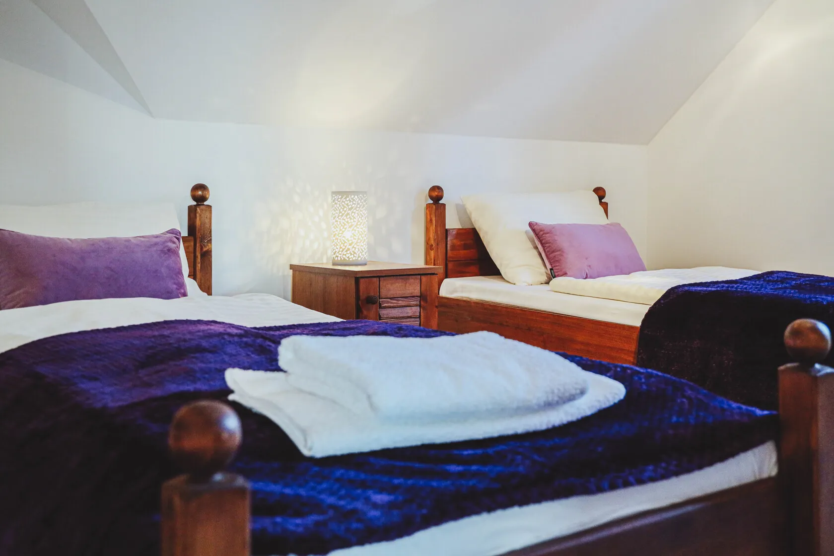 Izba s dvoma jednolôžkovými posteľami s bielym a fialovým posteľným prádlo, stolíkom a rozsvietenou lampou medzi posteľami.
