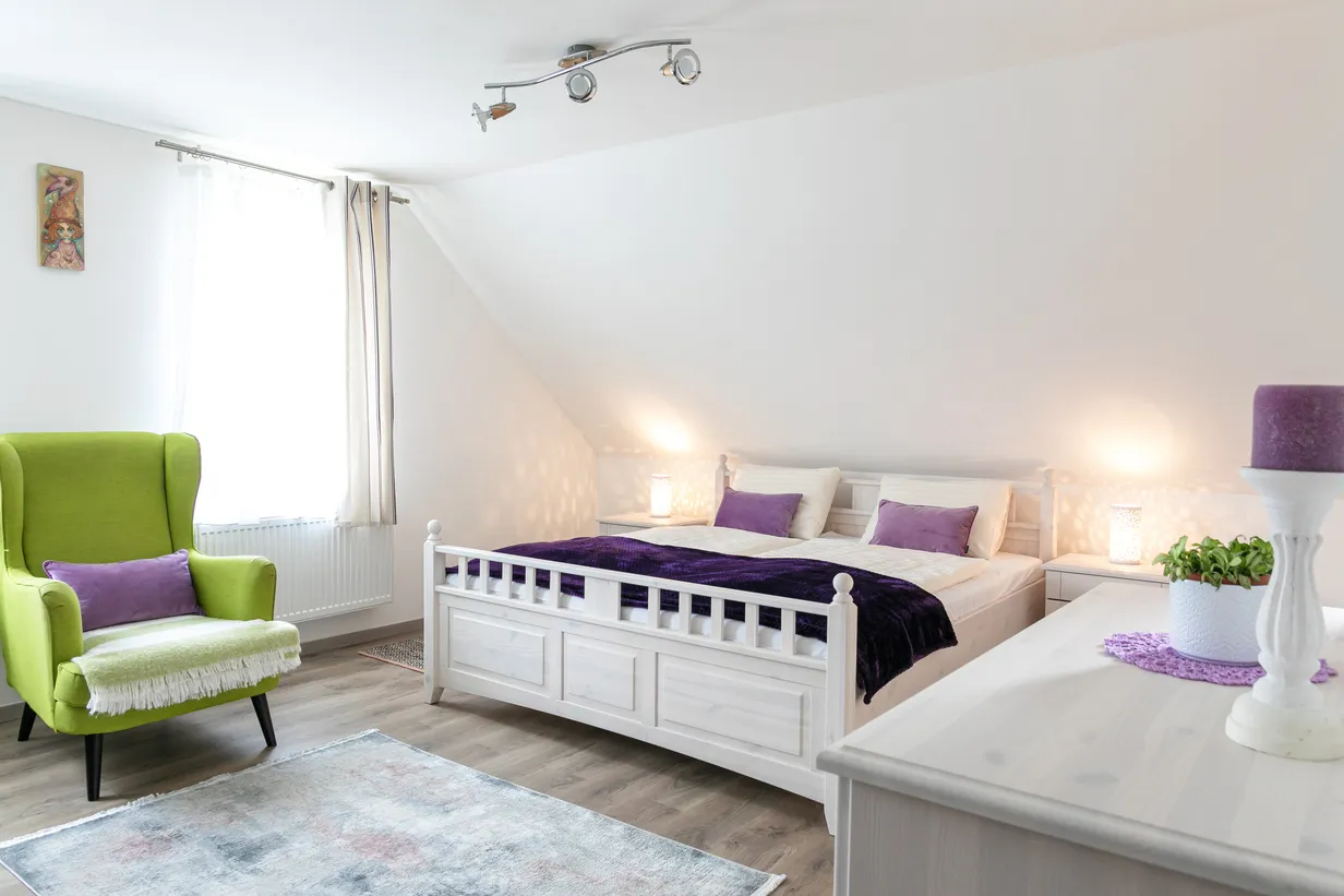 Svetlá spálňa so zeleným kreslom, bielou posteľou s fialovými vankúšmi a fialovou prikrývkou, nočnými stolíkmi a dekoráciou s fialovou sviečkou.