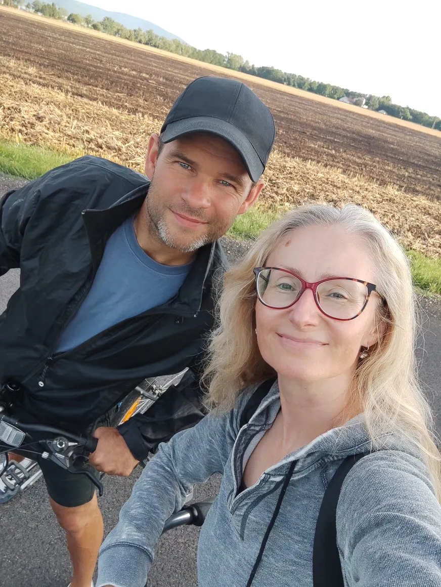 Muž v čiernej bunde a čiernej šiltovke a žena v sivom pulóvri s okuliarmi stoja vedľa bicyklov na ceste pri poli.