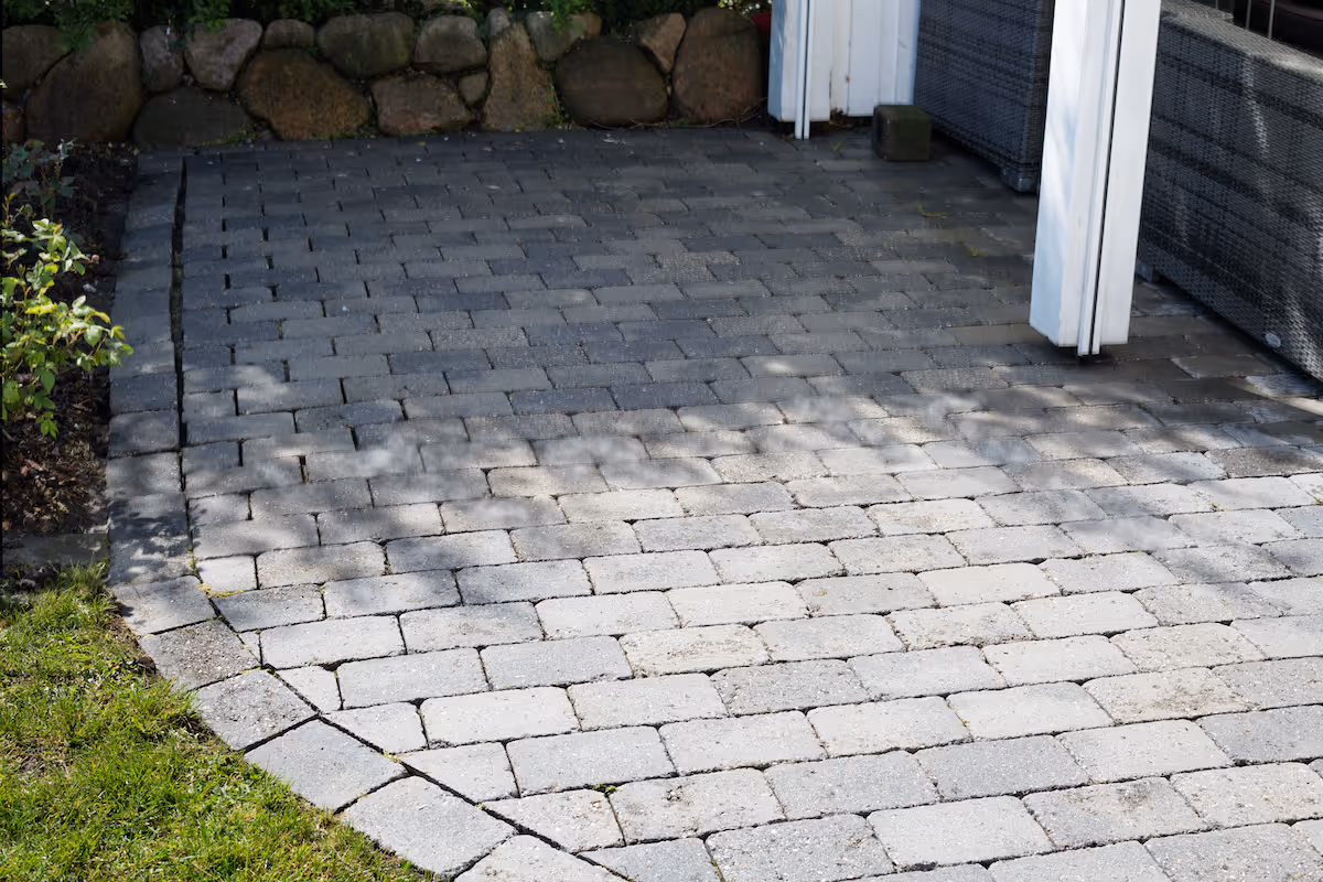 Flisebelagt terrasse med skygger, omgivet af græs og en stenmuret havekant.