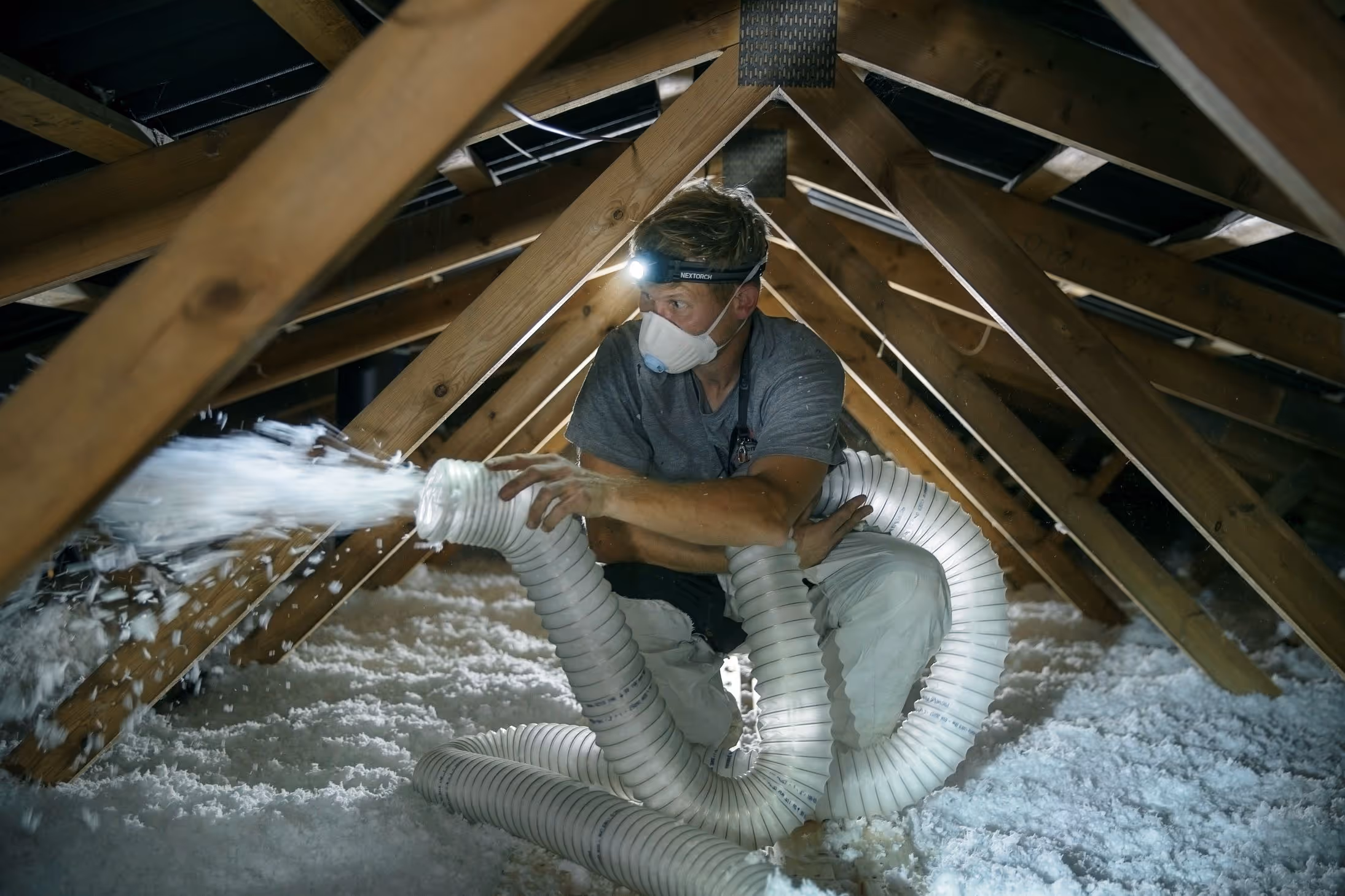 Mand iført ansigtsmaske og pandelampe sprøjter isoleringsmateriale i en loftsrum med træspær.