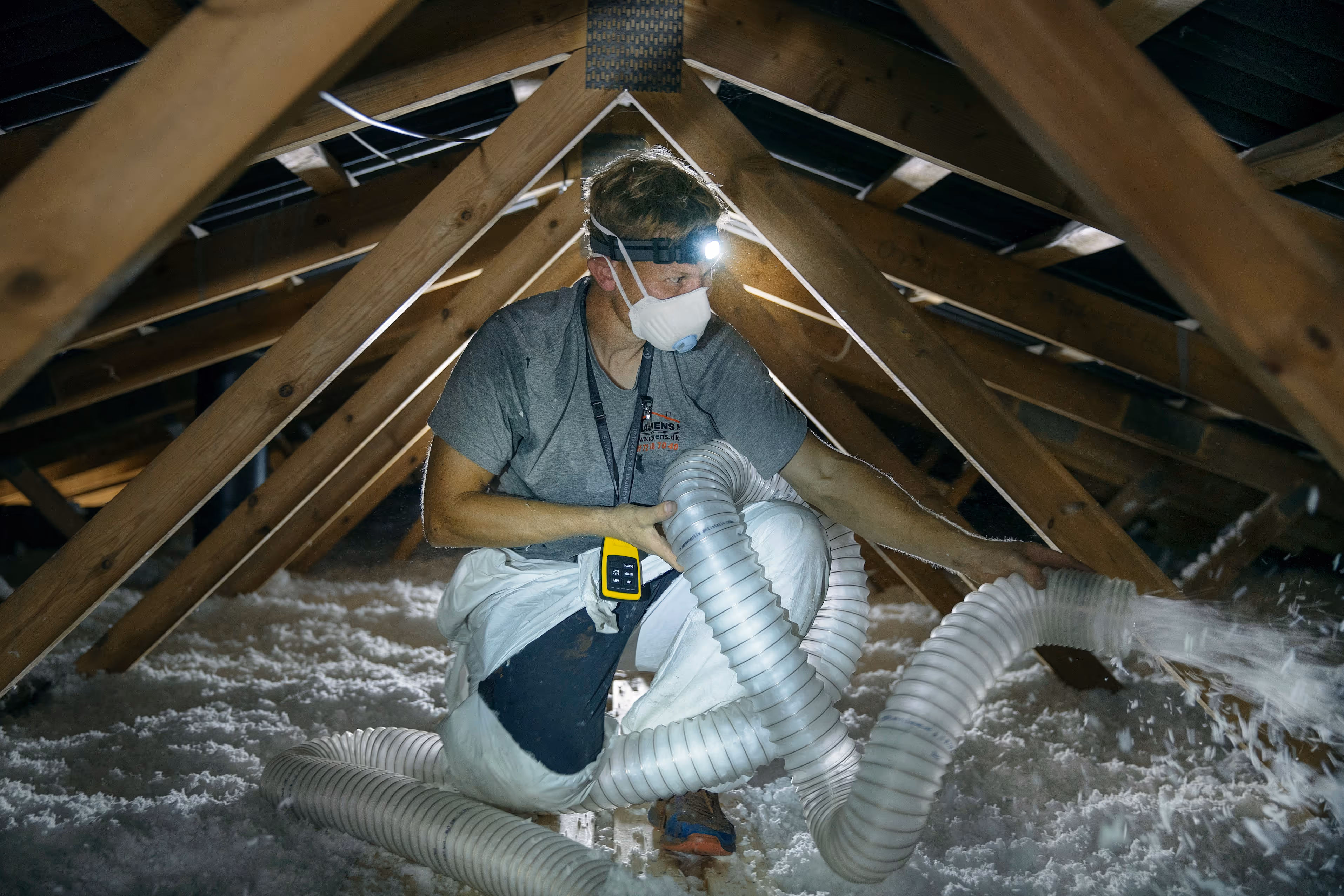 En arbejder med maske og pandelygte isolerer et loft ved at blæse hvid isoleringsuld gennem en stor slange.