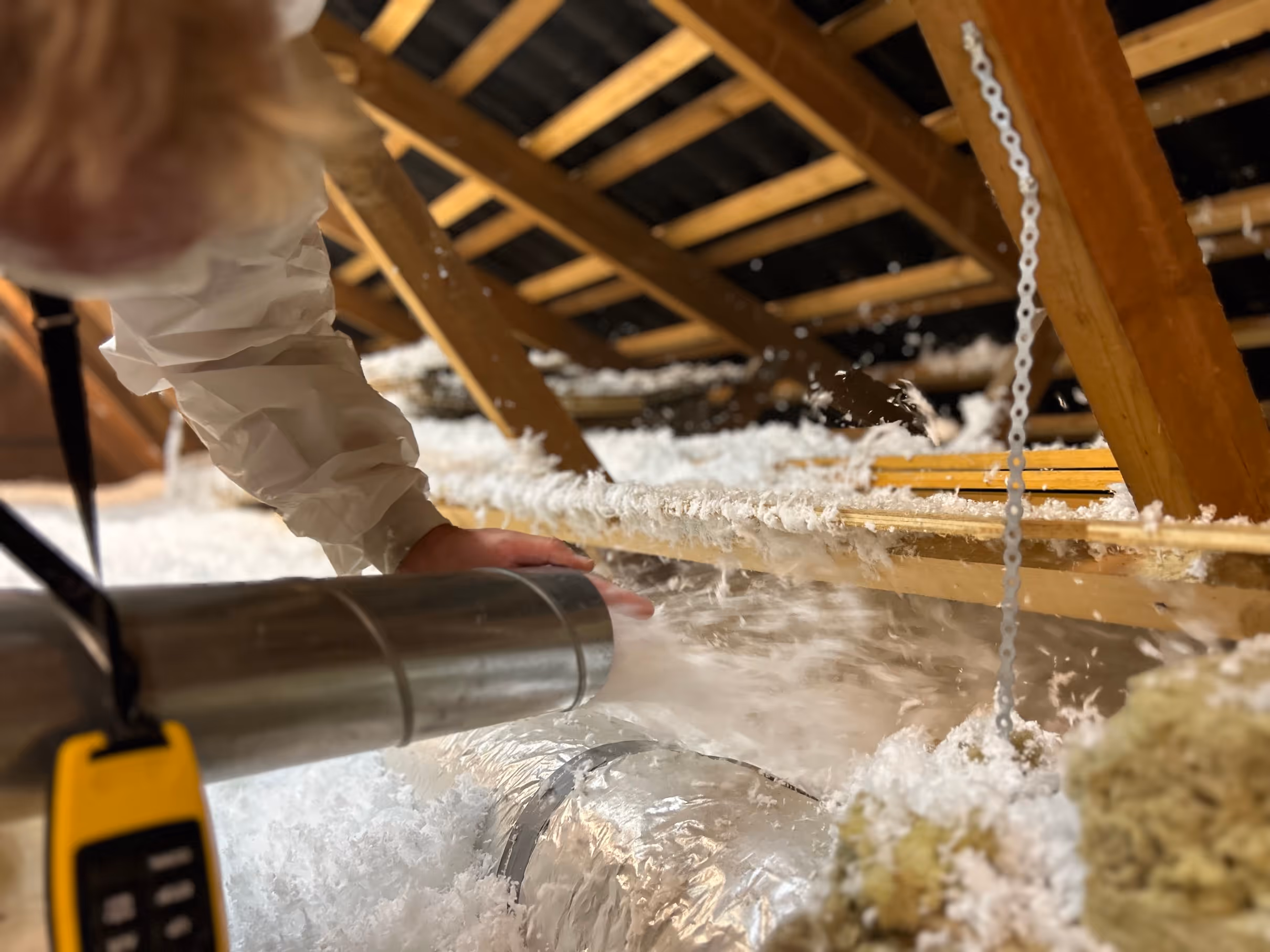 En person i beskyttelsesdragt blæser hvid isoleringsmateriale ind på loftet i en tagkonstruktion.