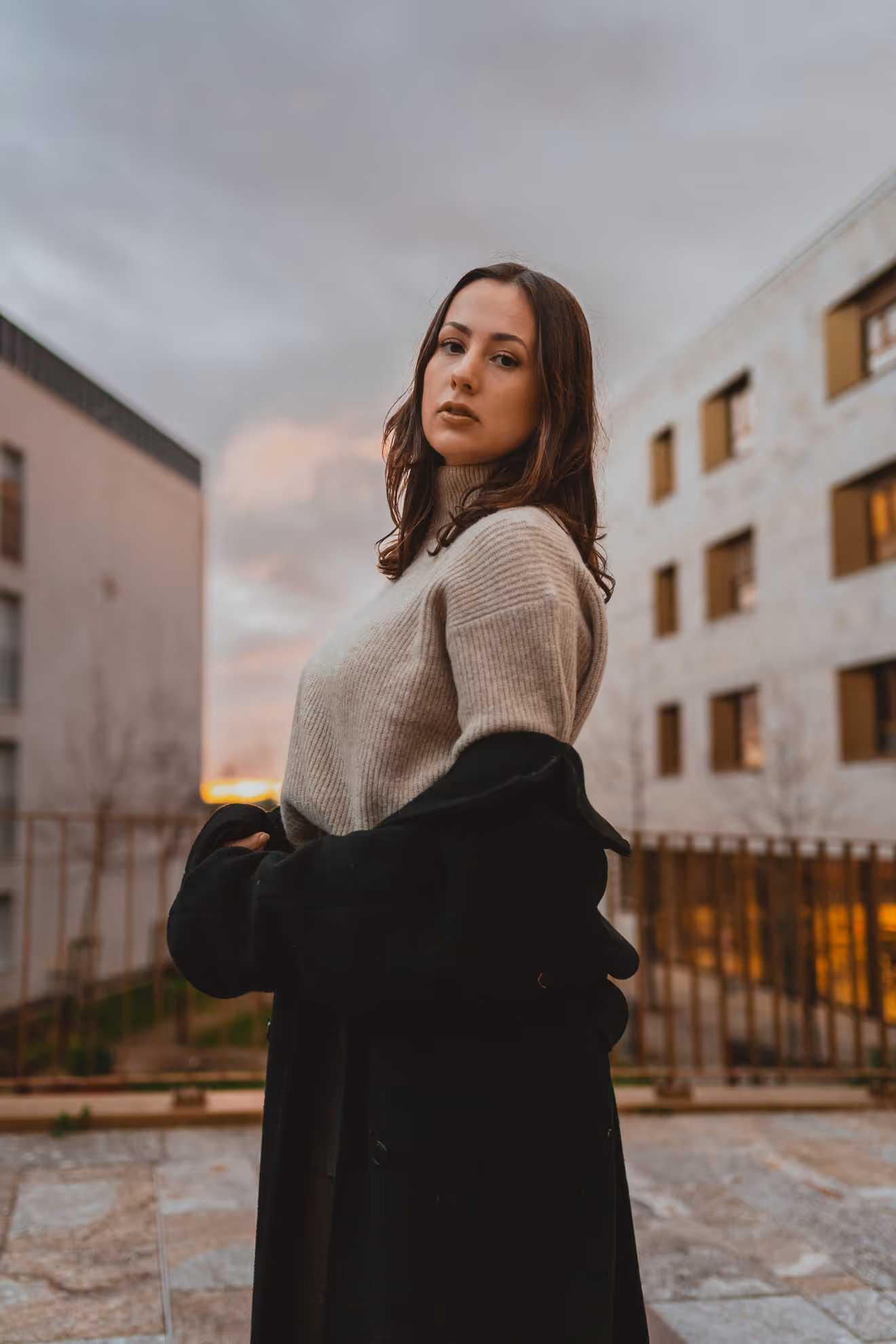 Jeune femme regardant la caméra, portant un pull en laine beige et un manteau noir, avec des bâtiments flous en arrière-plan au coucher du soleil.