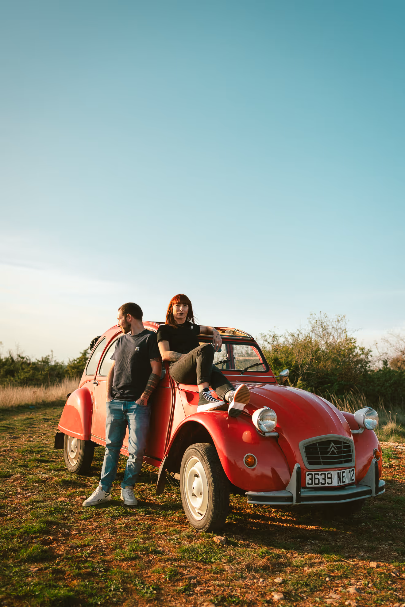 Un homme debout et une femme assise sur le capot d'une voiture ancienne rouge dans un champ sous un ciel bleu clair.