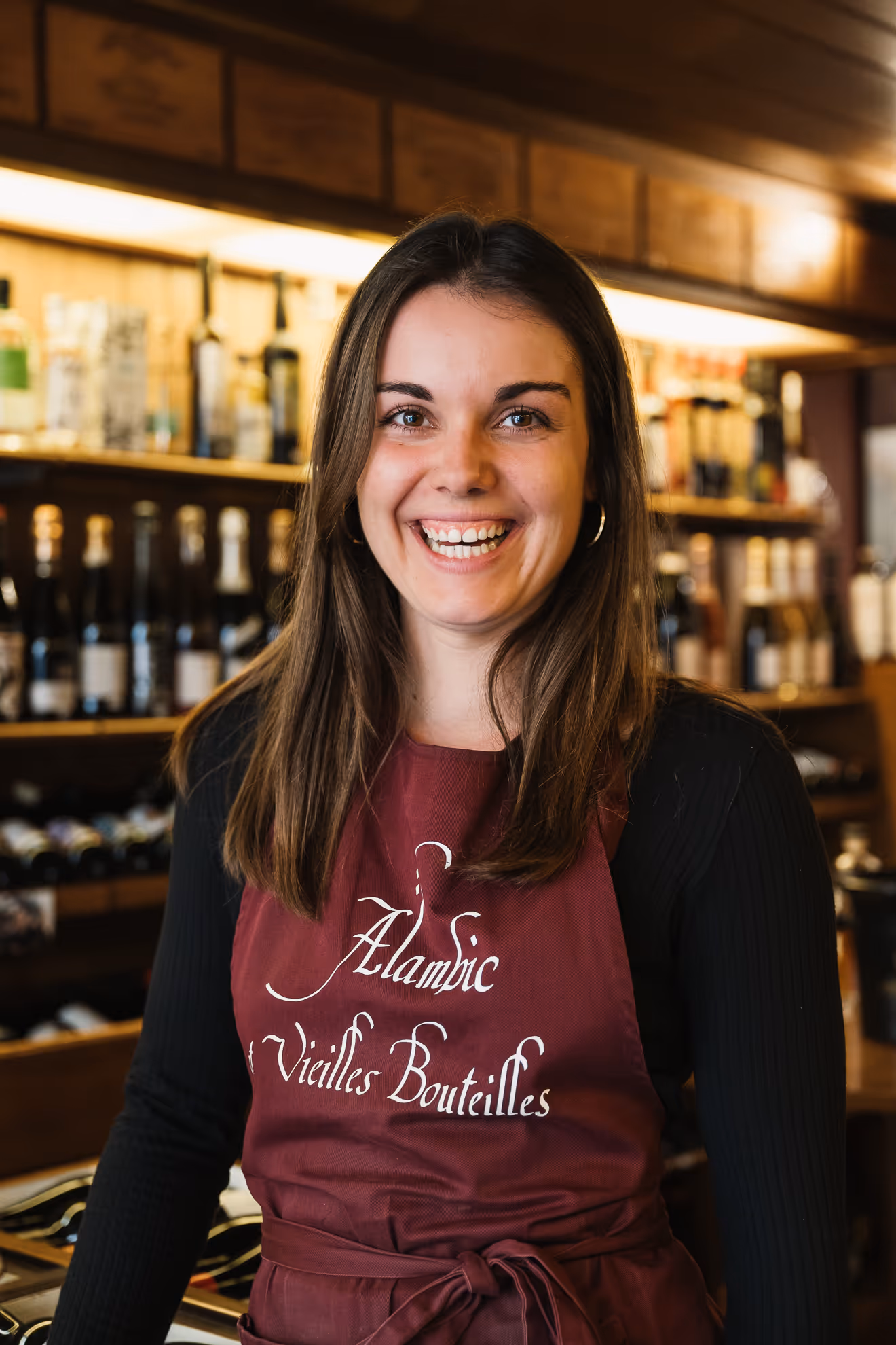 Femme souriante portant un tablier bordeaux avec l'inscription « Alambic et Vieilles Bouteilles », debout devant des étagères remplies de bouteilles.